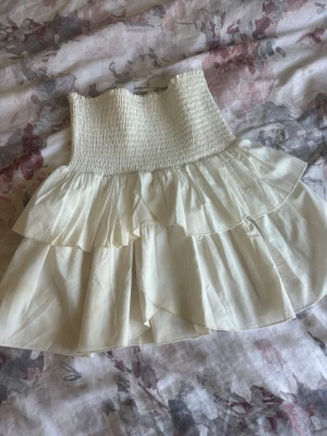 Vit volangkjol med smock - Supersöt kort kjol i vitt med bred smock i midjan och dubbla volanglager. Kjolen har en luftig och flowy känsla, perfekt för varma dagar. Tillverkad i ett tunt, mjukt material som ger en romantisk vibe. Finns lite små fläckar som syns på närahåll. Nypris 450