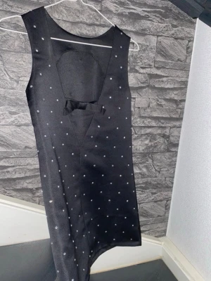 Svart prickig ärmlös klänning - Snygg svart ärmlös klänning med små diamanter och en dekorativ rosett på baksidan. Klänningen har en rak passform och är tillverkad i ett glansigt material som ger en elegant look. 