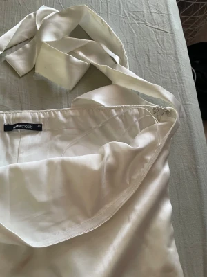 Vit omlottklänning från Gina Tricot - Säljer en elegant vit topp från Gina Tricot i blankt satintyg. Toppen har breda knytband vid halsen och dragkedja i sidan för enkel påklädning. Perfekt för dig som gillar stilrena och trendiga plagg.