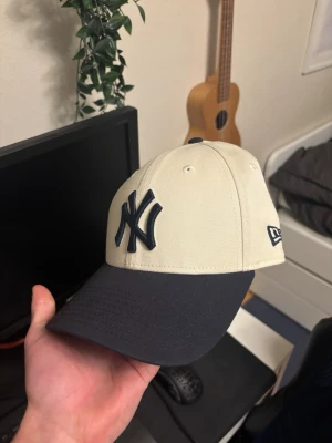  New Era Yankees keps - Snygg beige keps från New Era med klassisk New York Yankees-logga i svart framtill. Skärmen är svart och kepsen har broderade ventilationshål samt justerbar passform bak. Perfekt för dig som vill ha en stilren och sportig look.
