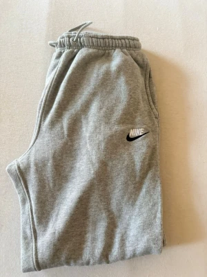 Gråa mjukisbyxor från Nike - Säljer ett par klassiska gråa mjukisbyxor från Nike med dragsko i midjan och resår vid bensluten. Byxorna har sidofickor och den ikoniska Nike-loggan broderad på ena benet. Perfekta för chill dagar eller träning. Knappast använda. 6 månader gamla men har endast legat i garderoben. Använda 2-3 gånger MAX.