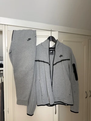 Grå Nike tech fleece komplett - Säljer ett grått set från Nike i storlek M. Setet består av en zip-hoodie med svarta detaljer och fickor samt matchande byxor med muddar. Båda delarna har Nike-logga broderad i svart. Perfekt för chill dagar hemma eller när du vill vara bekväm.