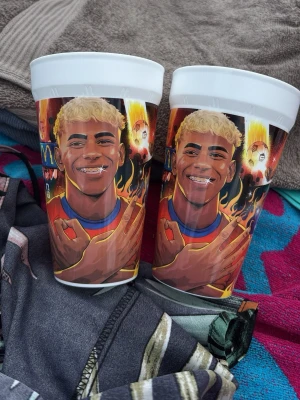 Två plastmuggar med färgglad tryck - Säljer två vita plastmuggar från mc med färgglad illustration av lamine yamal med blont hår och tandställning. Motivet har eldflammor och tecknade detaljer i bakgrunden. Perfekt för dryck på picknick eller hemma.