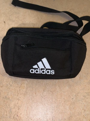 Adidas Svart magväska  - Vill köpa hörlurar därför väljer och sälja den totalt 2 fickor Snygg svart magväska från Adidas med vit logga framtill. Väskan har två dragkedjefack och justerbart band så du kan bära den runt midjan eller över axeln. Tillverkad i slitstarkt tygmaterial, perfekt för att ha med det viktigaste.