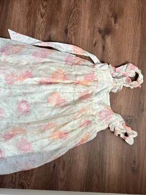 Blommig sommarklänning med volang - Söt kortklänning med blommigt mönster i pastellfärger som rosa och orange på vit botten. Klänningen har volangdetaljer vid axelbanden och knytband i midjan. Perfekt för varma dagar och har en luftig, romantisk känsla.