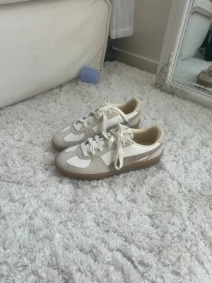 Vita och beige sneakers från Puma - Snygga sneakers från Puma i vitt och beige med klassisk retrodesign. Skorna har ovandel i mocka och textil, vita snören och en brun sula. Perfekta för dig som gillar stilrena och tidlösa sneakers.