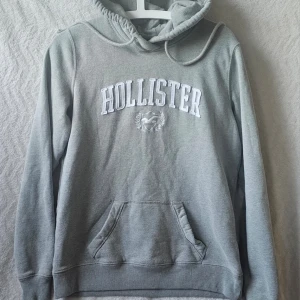 Grå hoodie från Hollister - Säljer en grå hoodie från Hollister med broderad logga och text på bröstet. Hoodien har känguruficka, dragsko i huvan och ribbade muddar. Perfekt för chill dagar eller när du vill ha en enkel och snygg look.
