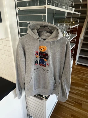 Ralph Lauren Hoodie - Säljer en grå Ralph Lauren hoodie med huva och dragsnören. På bröstet finns ett stort tryck av en björn i stickad tröja och jeans, samt texten 'Polo Bear by Ralph Lauren'. Hoodien har magficka och ribbade muddar. Perfekt för en chill och avslappnad stil.