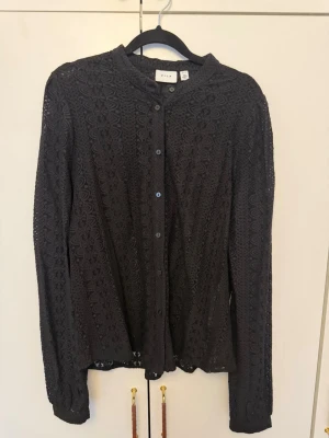 Svart spetsblus från VILA - Säljer en svart långärmad skjorta från VILA i genombruten spets med knappar framtill. Skjortan har en rundad krage och ett elegant mönster i tyget som ger en unik look. Perfekt för dig som vill ha något extra till outfiten.