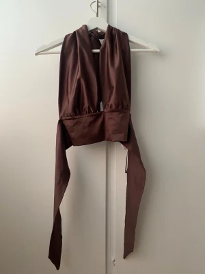 Brun omlott topp med knyt - Snygg brun omlott topp i satinliknande material med breda band som knyts i nacken och midjan. Toppen är ärmlös och croppad, perfekt för en trendig och stilren look.