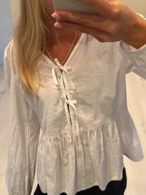 Vit blus med rosetter  - Säljer en söt vit blus med v-ringning och knytband framtill. Blusen har lång ärm och en lätt rynkad midja som ger en avslappnad och luftig känsla. Perfekt för dig som vill ha en stilren blus som passar till det mesta. Storlek 158/164