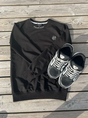 Svart sweatshirt från Philipp Plein - Svart sweatshirt från Philipp Plein med rund halsringning och ribbade muddar. Liten logga framtill på bröstet och stor vit logotyp på ryggen. Perfekt för en streetwear-look och enkel att matcha med jeans eller sneakers.