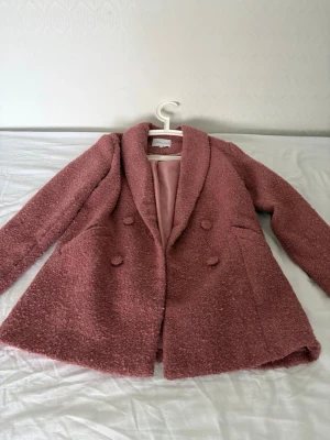 Rosa teddyjacka med dubbelknäppning - Supermysig teddyjacka i dammig rosa färg med dubbelknäppning och bred krage. Jackan har två fickor framtill och är fodrad med slätt tyg. Perfekt för att hålla sig varm och snygg under kyliga dagar.