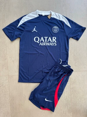 PSG  träningställ mörkblåt - Snyggt PSG matchställ med tröja och shorts i mörkblått med vita och röda detaljer. Tröjan har vita partier vid axlarna och tryck med Qatar Airways samt PSG-logga. Shortsen har röd detalj och PSG-emblem. Perfekt för fotbollsträning eller match.