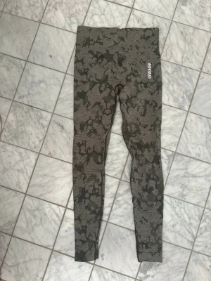 Camo-mönstrade leggings från Gymshark - Snygga camo-mönstrade leggings från Gymshark i grå och svart nyans. Hög midja och tight passform som framhäver benen. Perfekta för träning eller chill. Stretchigt material för maximal komfort.