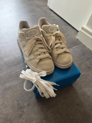 Beige Adidas sneakers med mockadetaljer - Snygga beige sneakers från Adidas med klassiska vita ränder på sidorna och logga på plösen. Skorna är i mocka och har en låg siluett. Extra vita skosnören medföljer. Perfekta för dig som gillar stilrena och neutrala färger.