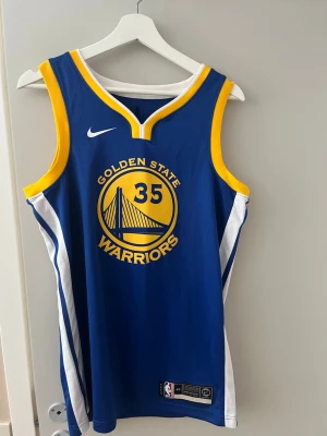 Golden State Warriors basketlinne - Blått basketlinne från Nike med Golden State Warriors-logga och nummer 35 på framsidan. Gula och vita detaljer längs kanterna och sidorna. På baksidan står det Durant och nummer 35. Perfekt för basketfans och träning.