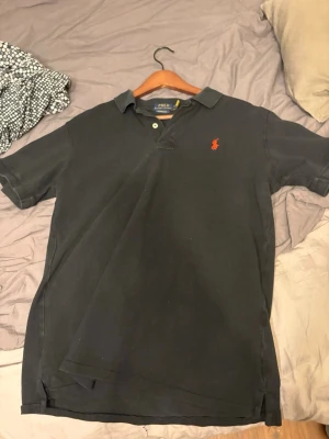 Svart pikétröja från Polo Ralph Lauren - Klassisk svart pikétröja från Polo Ralph Lauren med röd broderad logga på bröstet. Tröjan har krage, knappar vid halsen och korta ärmar. Perfekt till en avslappnad men ändå stilren look.