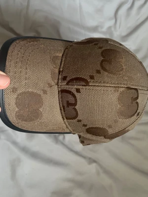 Brun Gcci keps med monogrammönster - Säljer en brun Gcci keps med mörkbrunt monogrammönster och svart kant på skärmen. Kepsen har en klassisk form och är tillverkad i ett tåligt tygmaterial. Perfekt för dig som vill ha en stilren accessoar med unik design.