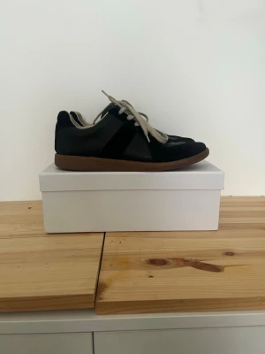 Svarta masion margiela gats  - Stilrena svarta sneakers från maison margiela med beige skosnören och bruna gummisulor. Skorna har en låg profil och är tillverkade i en mix av mocka och läder, vilket ger en snygg kontrast i material och textur. Perfekta för dig som gillar minimalistisk design.