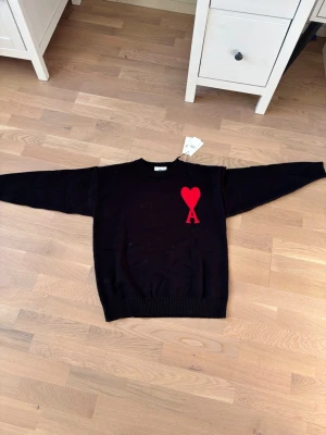Svart stickad tröja från Ami Paris - Svart stickad tröja från Ami Paris med röd hjärtlogga och bokstaven A på bröstet. Tröjan har rund halsringning, ribbade muddar och är gjord i mjukt material. Perfekt för dig som gillar stilrena plagg med en twist.