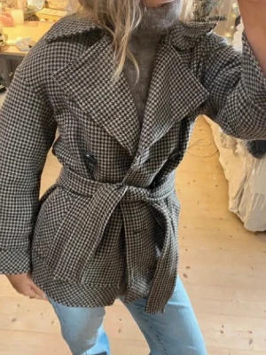 Rutig kappa med bälte Zara  - Snygg rutig kappa i svart, vit och brun med breda slag och knytskärp i midjan. Dubbelknäppt framtill med stora svarta knappar och två fickor. Klassisk modell som ger en cool och trendig look. Perfekt till höst och vinter.