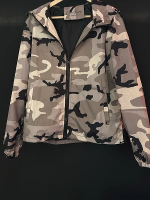 Camo vindjacka från Lindfors - Snygg vindjacka från Lindfors med grått, svart och beige camouflagemönster. Jackan har dragkedja framtill, huva och två sidofickor. Perfekt för dig som gillar streetwear och vill ha en lätt jacka med coolt mönster.