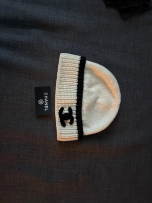 Vit och svart mössa från Chanel - Snygg vit stickad mössa från Chanel med svart kant och den klassiska Chanel-loggan broderad framtill. Mössan har en ribbstickad uppvikt kant och är tillverkad i mjukt material som känns lyxigt och trendigt.
