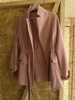 Ljusrosa parkajacka från Blanka - Säljer en ljusrosa parkajacka från Blanka med dragsko i midjan och stora fickor framtill. Jackan har långa ärmar med knappdetalj vid mudden och en avslappnad passform. Perfekt för vår och höst.