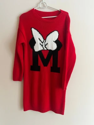 Röd stickad tröja med M och rosett - Säljer en röd stickad tröja med ett stort svart M och en vit rosett framtill. Tröjan har långa ärmar, rund halsringning och ribbade muddar. Perfekt för dig som gillar färgglada och lekfulla plagg.