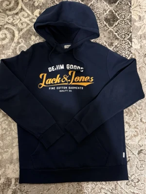 Marinblå hoodie från Jack & Jones - Snygg marinblå hoodie från Jack & Jones med trycket 'Denim Goods' och 'Fine Cotton Garments' i vitt och gult på bröstet. Klassisk huva med dragsko och mjukt bomullsmaterial. Perfekt för en avslappnad streetwear-look.