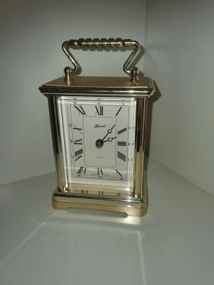 Klassisk guldfärgad bordsur - Stilren guldfärgad bordsur från Kienzle med rektangulär form och dekorativt handtag upptill. Uret har romerska siffror på vit urtavla och detaljer i svart. Tillverkad i metall med blank finish, perfekt som elegant inredningsdetalj.