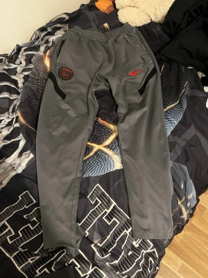 Gråa PSG x Nike mjukisbyxor - Snygga grå mjukisbyxor från Nike i samarbete med PSG. Byxorna har elastisk midja med dragsko, röda detaljer som Nike-logga och PSG-emblem på ena benet. Perfekta för en sportig och avslappnad stil.