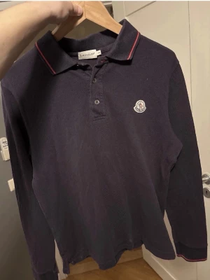 Mörkblå Moncler pikétröja - Snygg mörkblå långärmad pikétröja från Moncler med klassisk krage och två knappar. Tröjan har röda detaljer på kragen och ärmsluten samt Moncler-logga på bröstet. Perfekt för en stilren och avslappnad look. Inget kvitto 