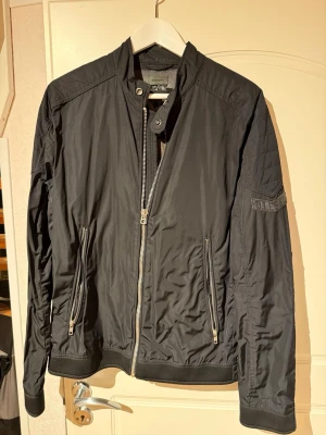 Svart bomberjacka från Diesel - Snygg svart bomberjacka från Diesel med raka linjer och coola detaljer. Jackan har två dragkedjeförsedda fickor framtill, ståkrage med tryckknapp och diskret logga på ärmen. Materialet är lätt och ger en modern look.