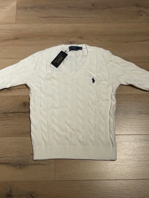 Vit kabelstickad tröja Polo Ralph Lauren - Vit kabelstickad tröja från Polo Ralph Lauren med V-ringning och broderad marinblå logga på bröstet. Tröjan har ribbstickade muddar och är tillverkad i mjukt material. Perfekt för en stilren och klassisk look.