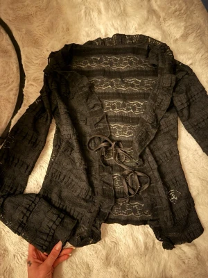 Svart spetsblus med volangkant - Spetsblus med volangkant längs urringningen och knytning framtill.