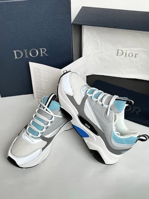 Dior vita och grå chunky sneakers - Snygga chunky sneakers från Dior i vitt, grått och svart med blå detaljer. Ovandelen är i mesh och läder med perforerade partier och coola kontrastsömmar. Tjock sula med svart och blå inslag. Vita och blå snören samt branding på sidan.