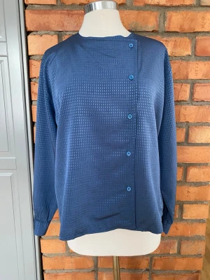 Blå asymmetrisk blus med knappar strl 44 - Stilren blå blus med diskret rutmönster och asymmetrisk knäppning framtill. Blusen har långa ärmar med knapp vid ärmslut och är tillverkad i ett glansigt, mjukt material som ger en elegant känsla. Perfekt för dig som gillar en unik twist på klassiska plagg.