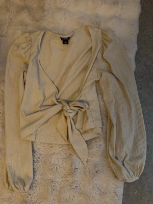 Beige omlottopp - Trendig beige omlottopp från Monki med långa puffärmar och knyt i midjan. Toppen är croppad och har en snygg v-ringning. 