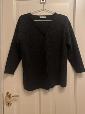 Svart kofta från Lord & Island - Mysig svart kofta från long Island med knäppning framtill och 3/4-ärmar. Koftan har en enkel, rak passform och är tillverkad i ett mjukt, lite luddigt material som känns skönt mot huden. Perfekt att slänga över en topp. St M 
