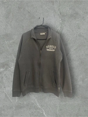 Grå zip hoodie från Dobber | L - Mörkgrå hoodie från Dobber med dragkedja framtill och två fickor. Tryck med 'Dobber 1974' på bröstet. Tillverkad i mjukt sweatshirtmaterial och har ribbade muddar vid ärmar och nederkant. Perfekt för en avslappnad stil.