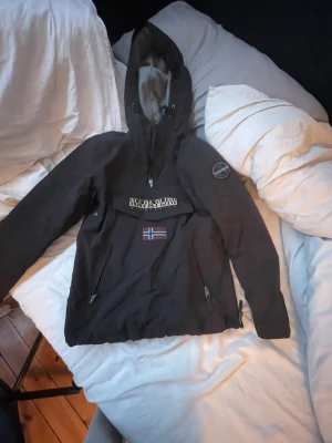 Brun vinterjacka  från Napapijri - Säljer en brun vinterjacka från Napapijri med stor huva och dragkedja framtill. Jackan har en stor ficka på magen med Napapijri-logga och norsk flagga. Materialet är slitstarkt och vattenavvisande, perfekt för blåsiga dagar. Snygg patch på ärmen ger extra stil.