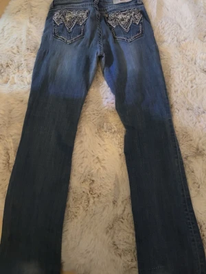 Blå bootcut jeans från Grace in La - Säljer ett par blå bootcut jeans från Grace in La med snygga broderade detaljer och stenar på bakfickorna. Jeansen har normal midja, klassisk femficksmodell och är gjorda i stretchigt denimtyg. Perfekta för dig som gillar en avslappnad men ändå trendig look.