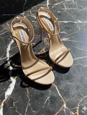 Beige stilettklackar från Steve Madden - Säljer ett par snygga beige pumps från Steve Madden med tunna remmar och hög stilettklack. Skorna har öppen tå och elegant design, perfekta för att lyfta vilken outfit som helst. Tillverkade i syntetmaterial med en stilren look.