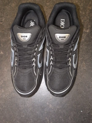 Svarta Dior B30 - Snygga svarta Dior B30 sneakers med reflekterande silverdetaljer och meshpaneler. Skorna har en chunky sula och tydlig Dior-logga på sidan. Perfekta för dig som vill ha en exklusiv och trendig look. Klassisk snörning och bekväm passform.