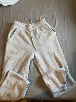 Ljusgrå mjukisbyxor med dragsko - Säljer ett par ljusgrå mjukisbyxor med resår i midjan och dragsko för skön passform. Byxorna har två sidofickor och muddar vid bensluten. Perfekta för chill dagar hemma eller när du vill vara bekväm. Tillverkade i mjukt sweatshirtmaterial.