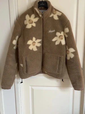 Brun teddyjacka med blommor från Vargal - Mysig brun teddyjacka från Vargal med stora vita och beige blommor över hela jackan. Jackan har hög krage, dragkedja framtill och två fickor med dragkedja. Perfekt för dig som vill ha en trendig och unik look i höst och vinter.