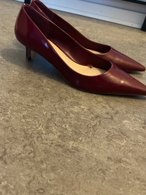 Vinröda pumps med låg klack från Mango - Snygga vinröda pumps från Mango med spetsig tå och låg klack. Skorna är i glansigt material och har en klassisk, elegant design som passar perfekt till både jeans och klänning. Perfekta för dig som gillar stilrena och trendiga skor.