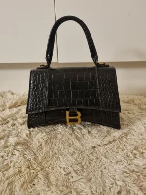 Svart Balenciaga krokodilväska - Snygg svart handväska från Balenciaga i krokodilpräglat läder. Väskan har ett topphandtag och en guldig B-detalj framtill. Stilren och trendig design med strukturerad form och exklusiv känsla. Perfekt accessoar för att lyfta din outfit.